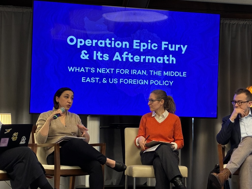 Elham Hoominfar sitting in a chair and speaking into a microphone on the stage with two other people and "Operation Epic Fury & Its Aftermath: What's Next for Iran, the Middle East, & US Foreign Policy" on a screen behind them