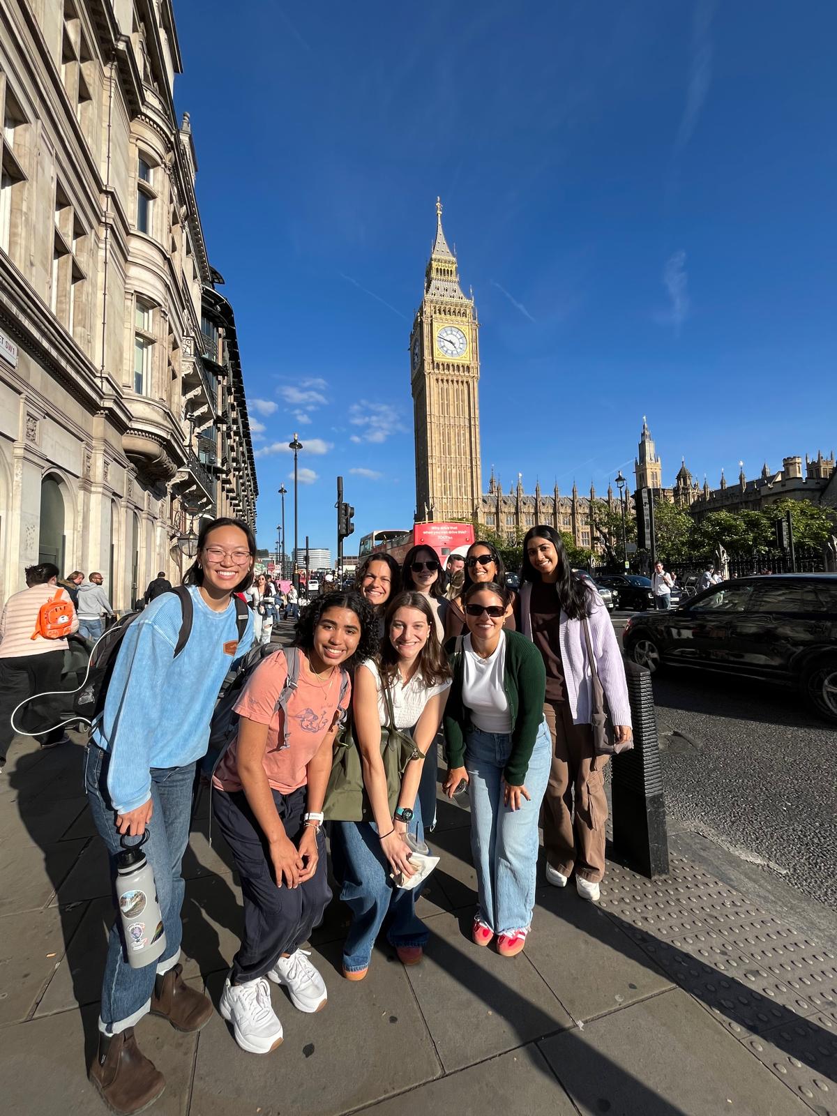 a group of young women standing in front of a large clock tower on a sunny day